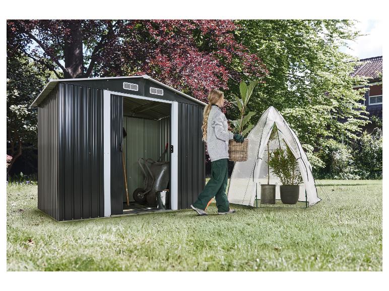 Vrouw draagt een plant naast een tuinhuisje en een kleine kas.