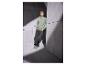 Man in groene sweater met 'Club 1990' en donkere jeans, leunend tegen een muur.