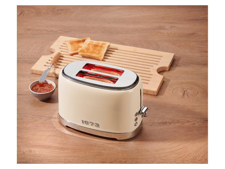 Crèmekleurige broodrooster met toast, jam en een houten snijplank op een houten tafel.