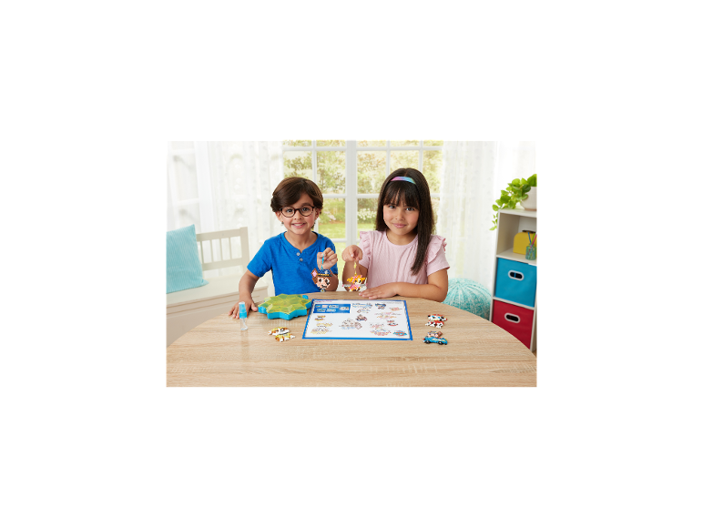 Twee lachende kinderen houden kleurrijke kralencreaties vast aan een houten tafel.