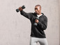 Man in zwarte hoodie en grijze broek, doet oefeningen met dumbbells.
