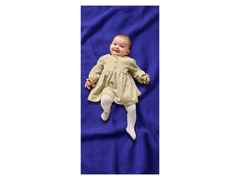 Blije baby in beige jurk en witte panty op een blauwe wafel deken.
