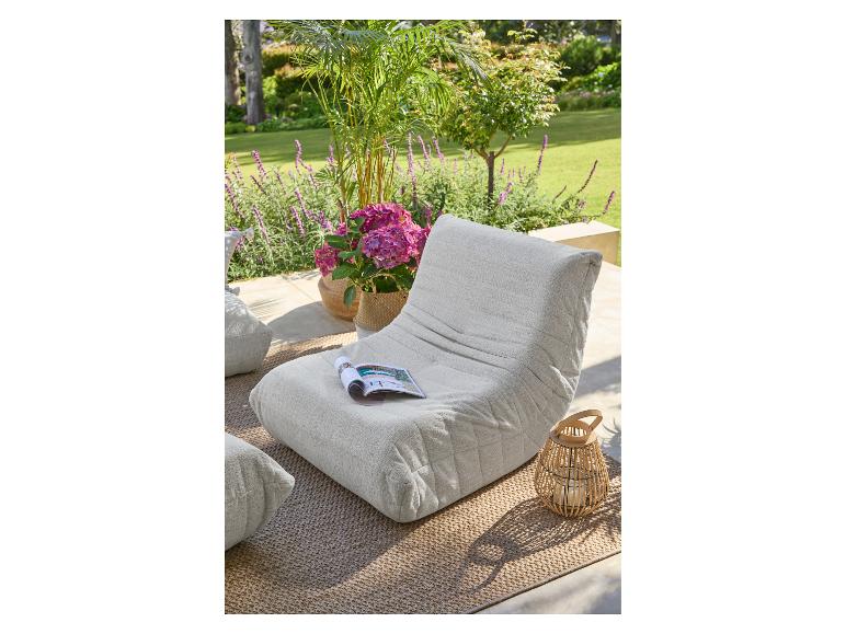 Beige buitenpoef met tijdschrift, planten en lantaarn op een vloerkleed.
