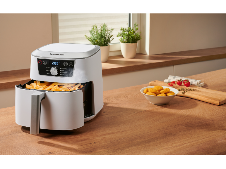 Witte airfryer met frietjes en een kom met kipnuggets op een houten tafel.