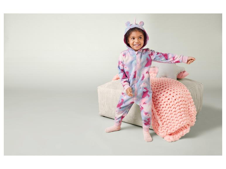 Een kind in een roze unicorn onesie staat op een bank.