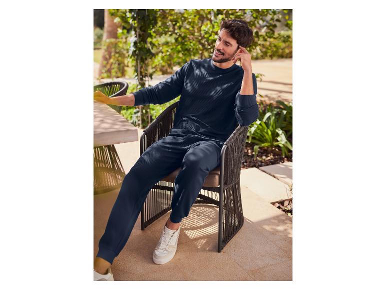 Man in een donkerblauwe sweater en broek, zittend op een stoel buiten.