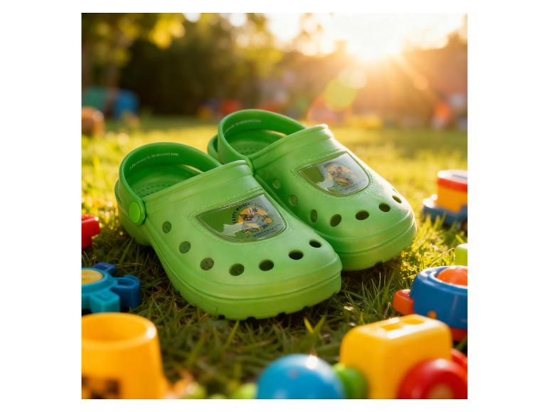 Groene kindersandalen met cartoonfiguren op gras met speelgoed bij zonsondergang.