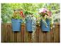 Drie blauwe plantenbakken met kleurrijke bloemen hangen aan een houten hek.