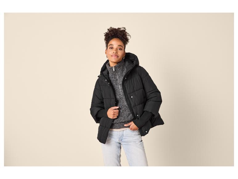Vrouw in zwarte pufferjas, grijze trui en lichte jeans.