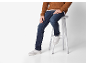 Man in bruine gebreide trui, blauwe jeans en witte sneakers, zittend op een kruk.