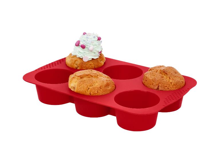 Rode siliconen bakvorm met zes holtes, waarvan drie muffins bevatten, één met lichtgroen glazuur.