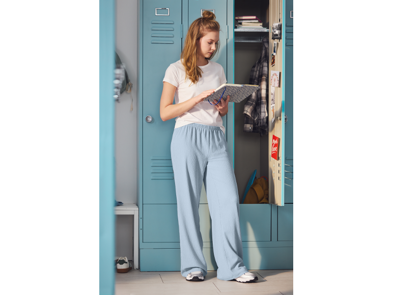 Meisje in wit t-shirt en lichtblauwe broek staat bij kluisjes, leest een notitieboekje.