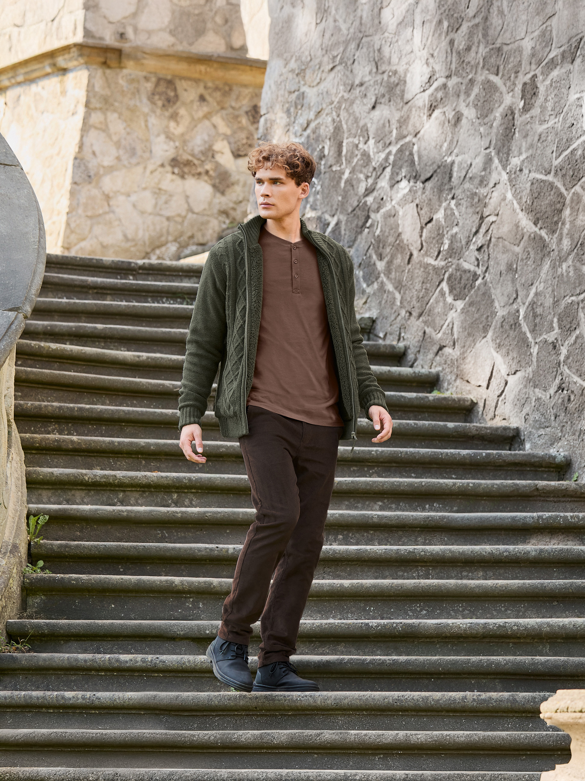 Man in groen gebreid vest, bruin T-shirt en bruine broek loopt stenen trap af.