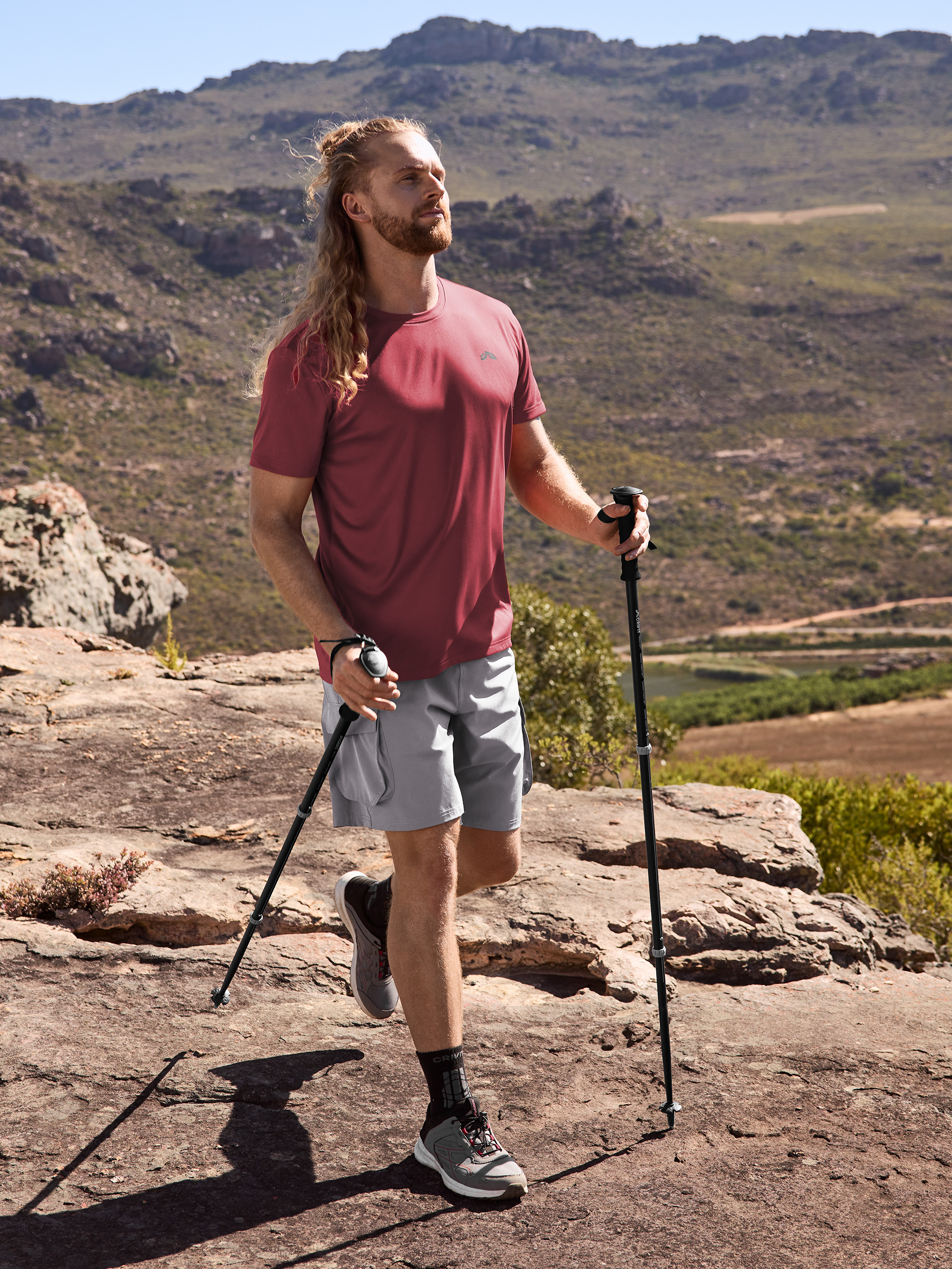 Man in wandel-T-shirt en korte broek, met trekkingstokken in bergachtig landschap.