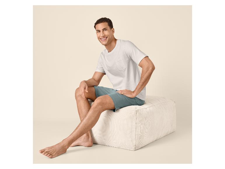 Lachende man in wit T-shirt en blauwe korte broek zittend op een poef.
