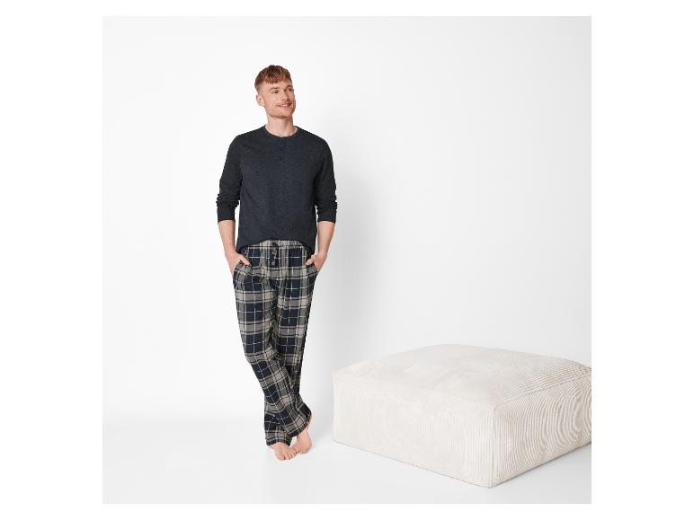 Man in geruite pyjama en grijs shirt, staand naast een beige poef.