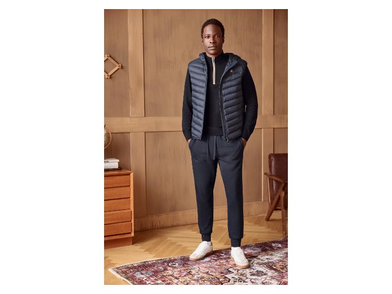 Man in marineblauw vest, zwarte trui en broek, witte sneakers, staand op een patroonkleed.