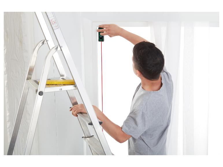 Een man meet een raam met een laserafstandsmeter, staand op een ladder.
