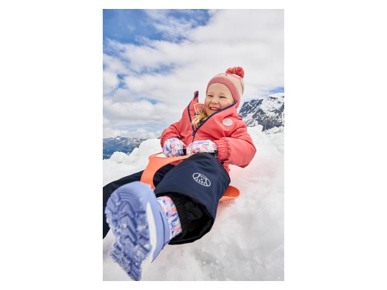 Lachend meisje in een roze jas en blauwe sneeuwbroek, zittend op een oranje slee in de sneeuw.