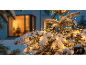 Sneeuwbedekte buitenkerstverlichting aan een boom, met een warm verlicht raam op de achtergrond.