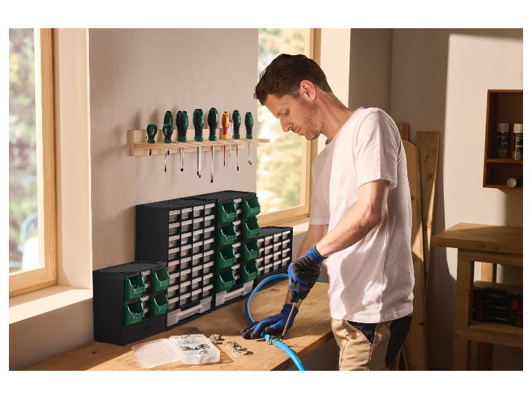 Man werkt met een schroevendraaier aan een slang op een werkbank met sorteerdozen en een schroevendraaierrek.