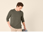 Man in donkergroen shirt met lange mouwen en beige broek, houdt een spijkerjack vast.