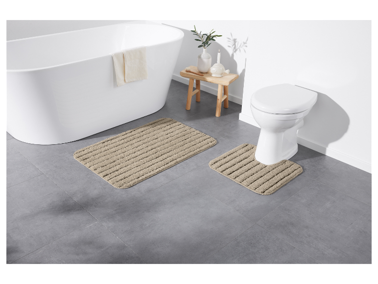 Badkamer met grijze tegels, wit bad en toilet, en beige badmatten.