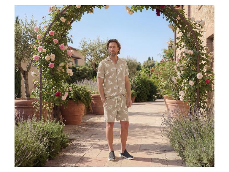 Man in een beige overhemd en korte broek met bladprint, in een tuin.