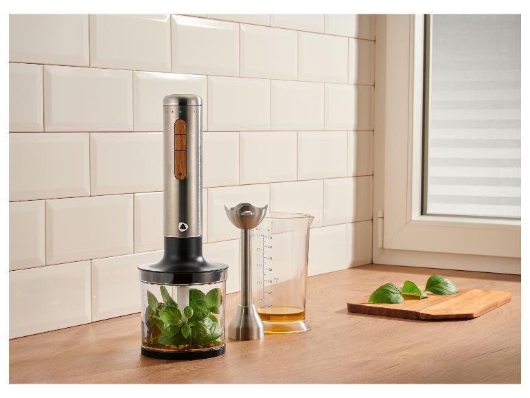 Staafmixer met basilicum in een bakje en maatbeker met olie op een houten aanrecht.