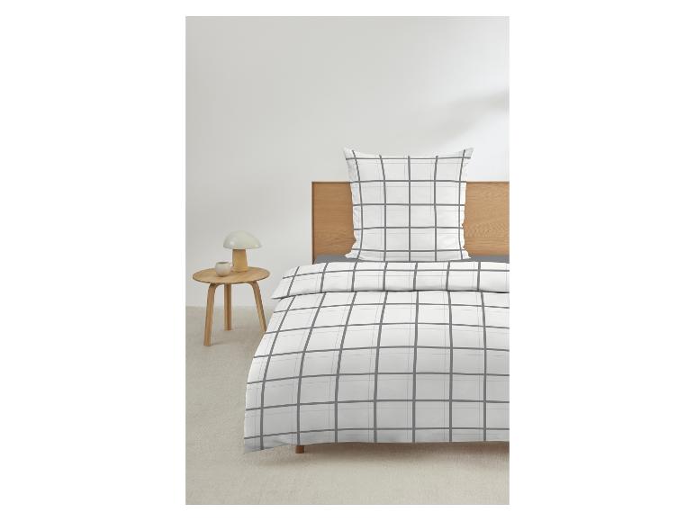 Wit beddengoed met grijs ruitpatroon op bed met houten hoofdbord, nachtkastje met lamp ernaast.