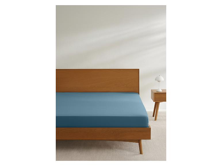 Bed met houten frame en blauw hoeslaken, naast een nachtkastje met lamp.
