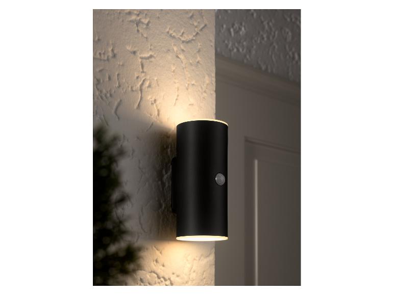 Een zwarte cilindrische buitenwandlamp met bewegingssensor verlicht een getextureerde muur.