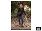 Vrouw in zwarte jas en jeans, met een fiets in een park.