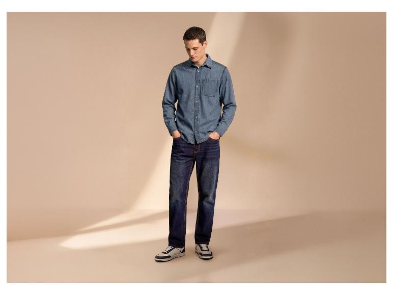 Man in spijkeroverhemd en jeans met handen in zakken, kijkt naar beneden.