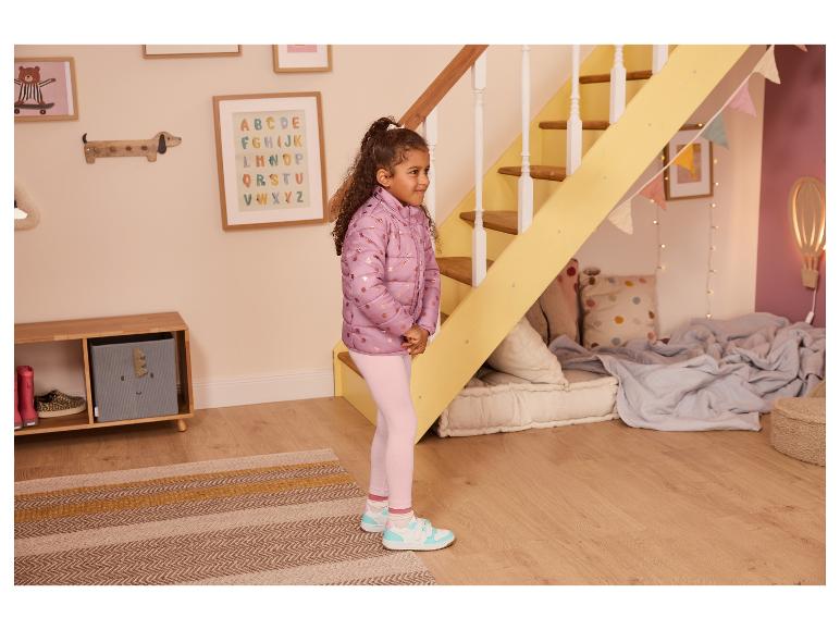 Meisje in paarse jas en roze legging staat in een kinderkamer met een trap.