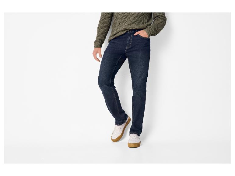 Man draagt een donkergroene gebreide trui, donkerblauwe jeans en witte sneakers met bruine zolen.