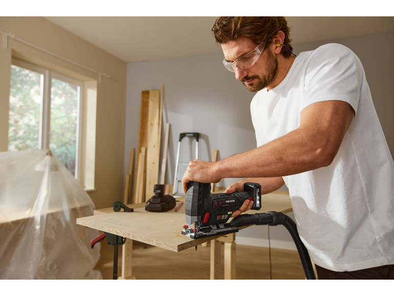 Man zaagt een houten plank met een decoupeerzaag, draagt veiligheidsbril en oordopjes.