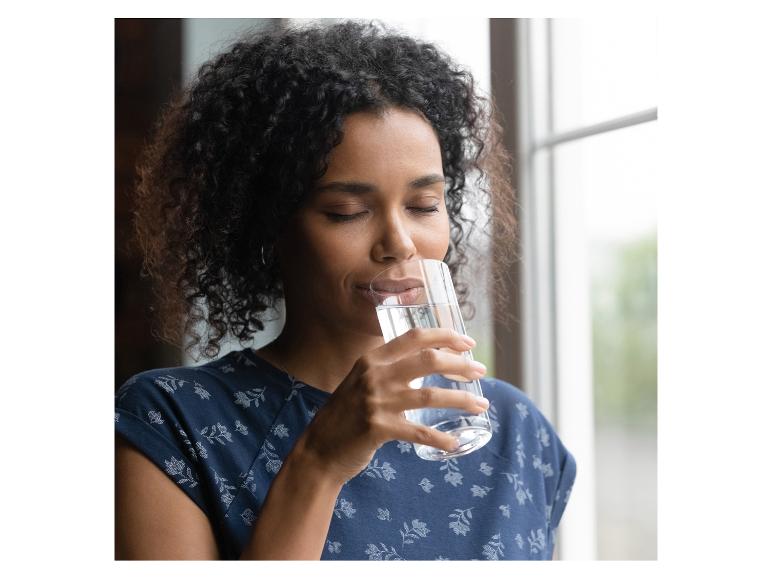 Vrouw met krullend haar drinkt water uit een glas, draagt een blauwe bloemenblouse.