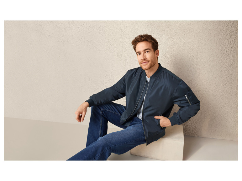 Een man draagt ​​een donkerblauwe bomberjack en blauwe jeans.