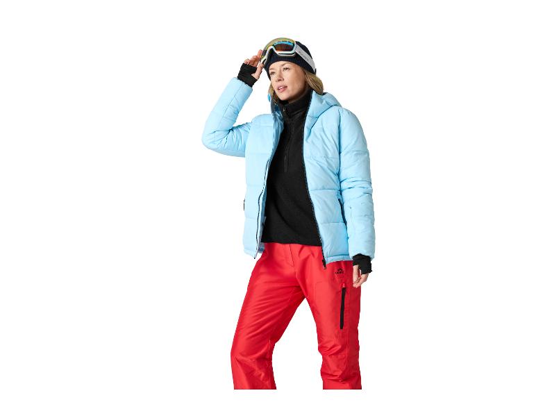 Vrouw in blauwe ski-jas, rode broek en skibril.
