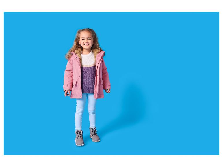 Lachend meisje in roze winterjas, witte en paarse trui, lichtblauwe jeans en grijze laarzen.