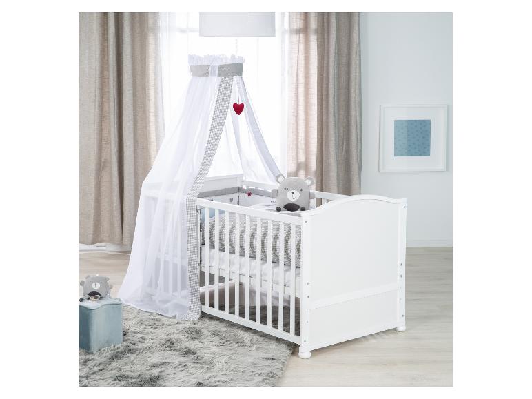 Wit babybedje met hemel, grijze pluche beer en vloerkleed in een kinderkamer.