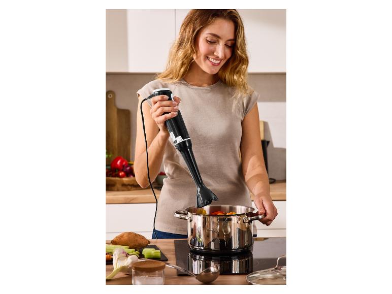 Vrouw gebruikt een staafmixer in een pan met groenten op een kookplaat.