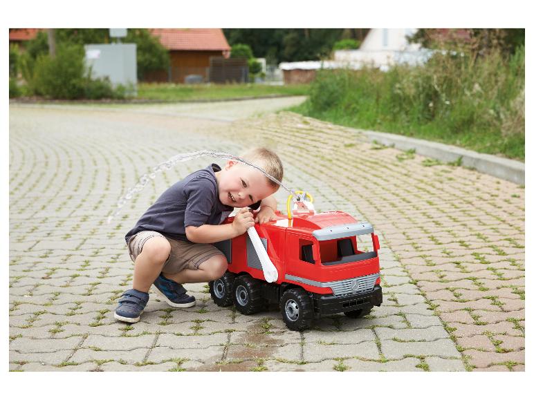 Lachende jongen speelt met een rode speelgoedbrandweerwagen die water spuit op een verharde ondergrond.