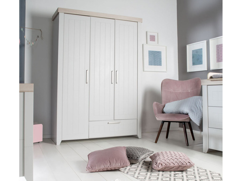 Een witte kledingkast met drie deuren en een lade, een roze stoel en decoratieve kussens.