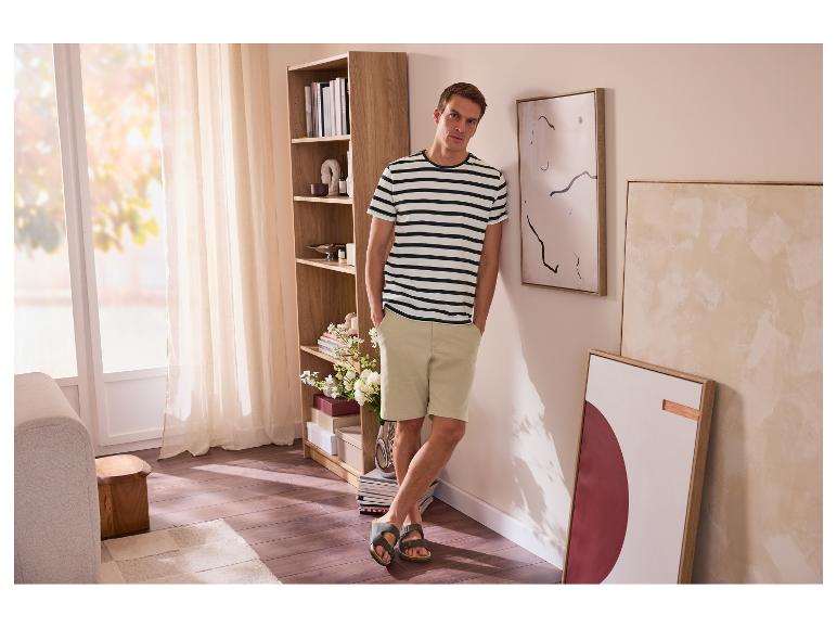 Man in gestreept T-shirt en korte broek, leunend tegen een muur in een woonkamer.