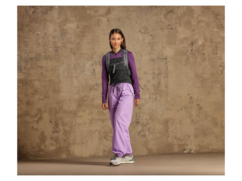 Een vrouw in paarse broek, een zwart vest en sneakers tegen een ruwe muur.