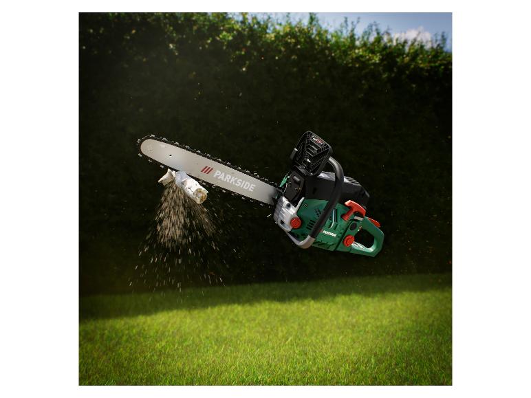 Kettingzaag zaagt een tak, met zaagsel in de lucht, tegen een groene heg en gras.