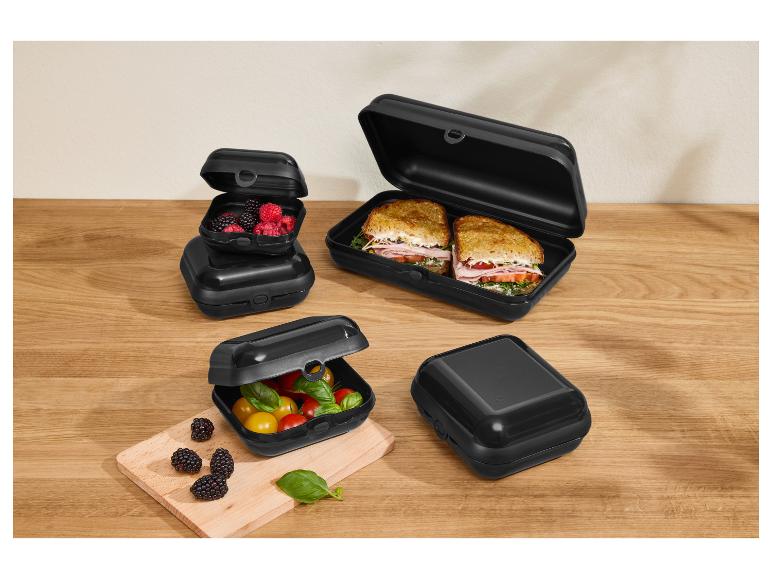 Zwarte lunchboxen met sandwiches, bessen en tomaten op een houten tafel.