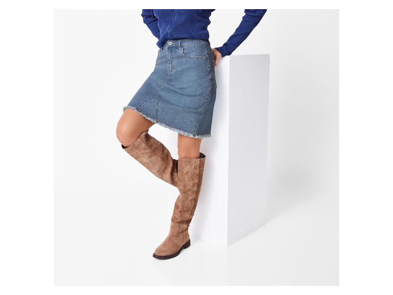 Vrouw in een blauwe trui en jeansrok, met hoge bruine laarzen.